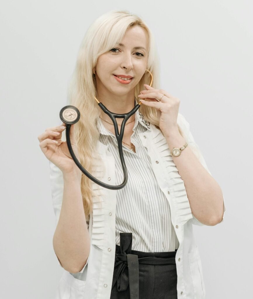 Blonde female doctor smiling while holding a stethoscope, wearing a white top on a plain background.